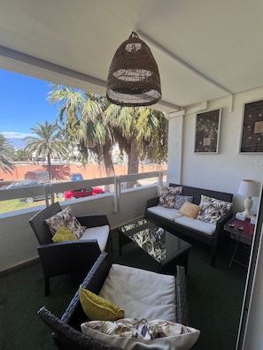 A balcony with wicker furniture, a hanging light fixture, and a view of palm trees and vehicles. - Lovely 2 Bedroom Apartment Near the Beach (Roquetas de Mar)