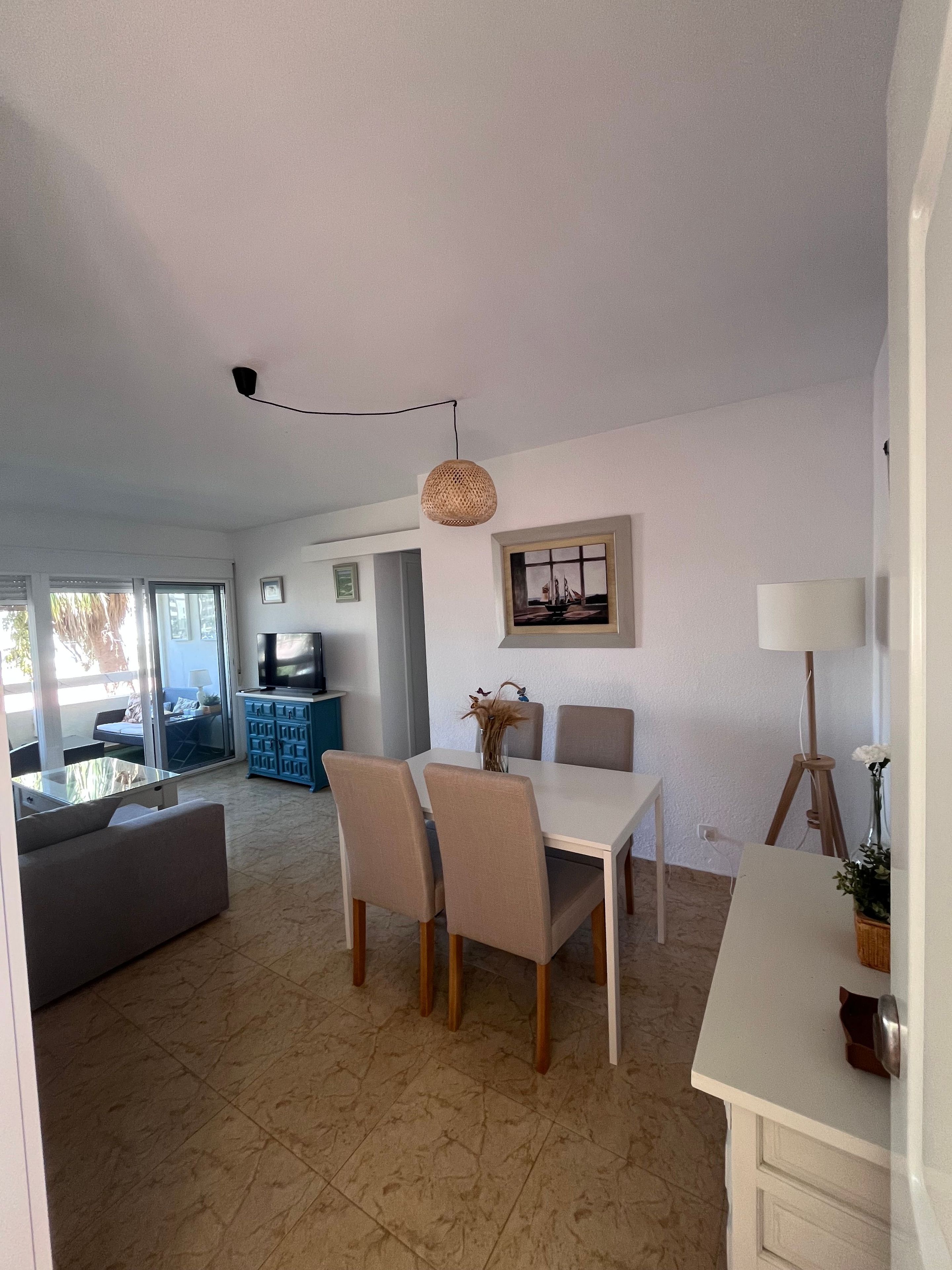 Un amplio salón con mesa de comedor, sillas y sofá. Hay un televisor y un jarrón decorativo sobre la mesa.