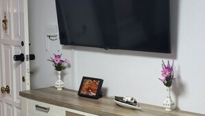 A white door with a brass handle, a white wall with a mounted television, and a wooden cabinet with drawers and a vase with flowers. - VAO Holiday Rental-los Narejos by Oasis (Los Alcazares)
