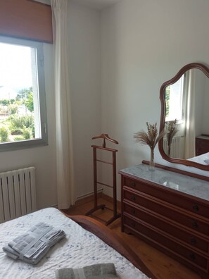 Ein Schlafzimmer mit Bett, Schminktisch, Spiegel, Heizkörper und Fenster mit Blick auf Grünflächen.