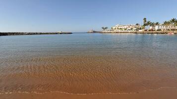 A beach with clear water, a sandy shore, and buildings in the background.