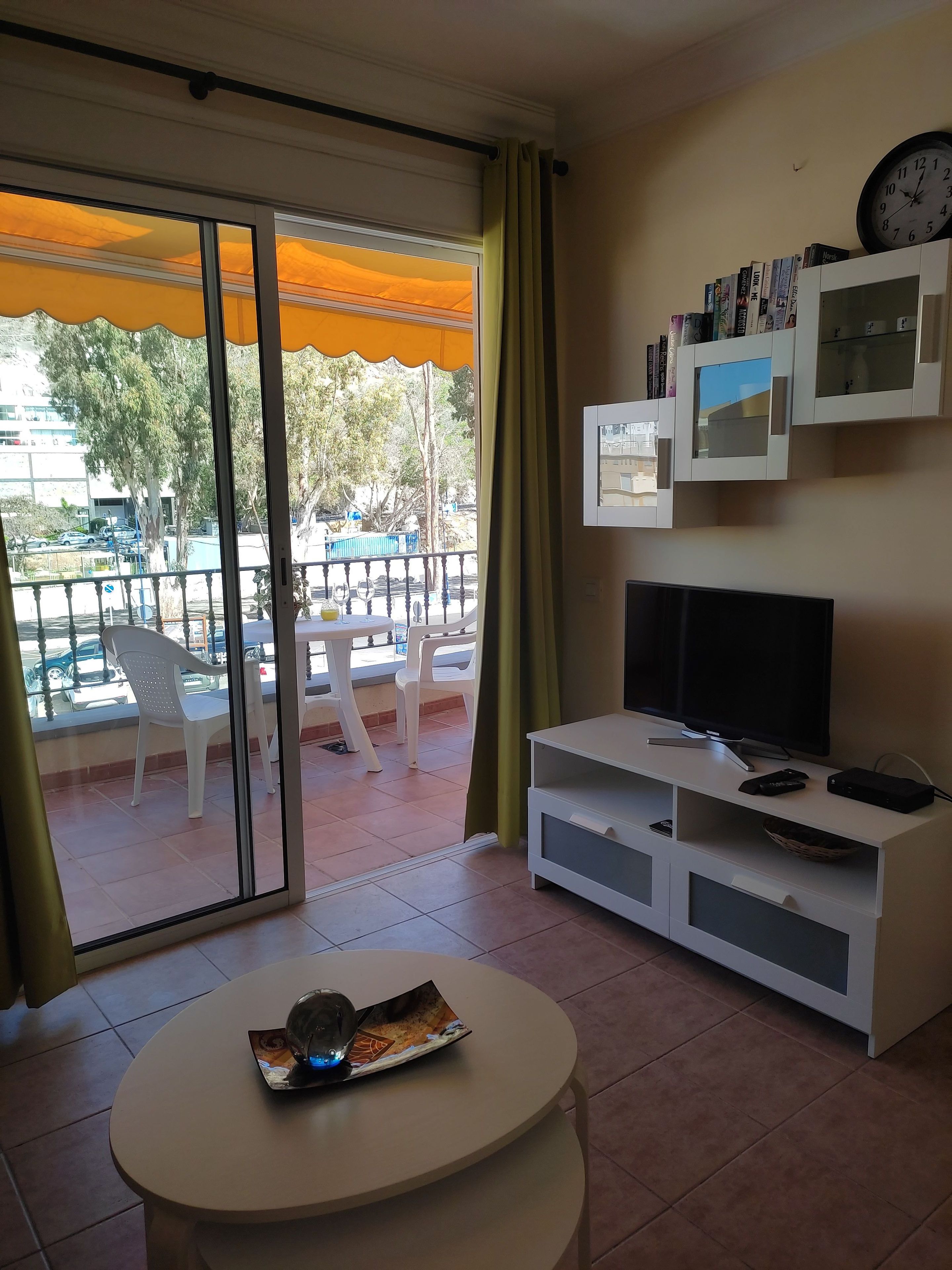 Una sala de estar con televisor, un mueble blanco para equipo audiovisual, una mesa redonda con una revista, y un balcón con sillas y una mesa.