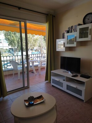 A living room with a TV, a white entertainment unit, a round table with a magazine, and a balcony with chairs and a table. - 1 Bedroom Apartment in the Center of Puerto de Mogan (Mogan)