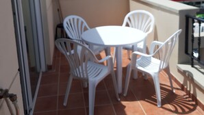 Una terrazza con sedie e tavolo di plastica bianca, pavimento piastrellato e vista su edifici sullo sfondo.