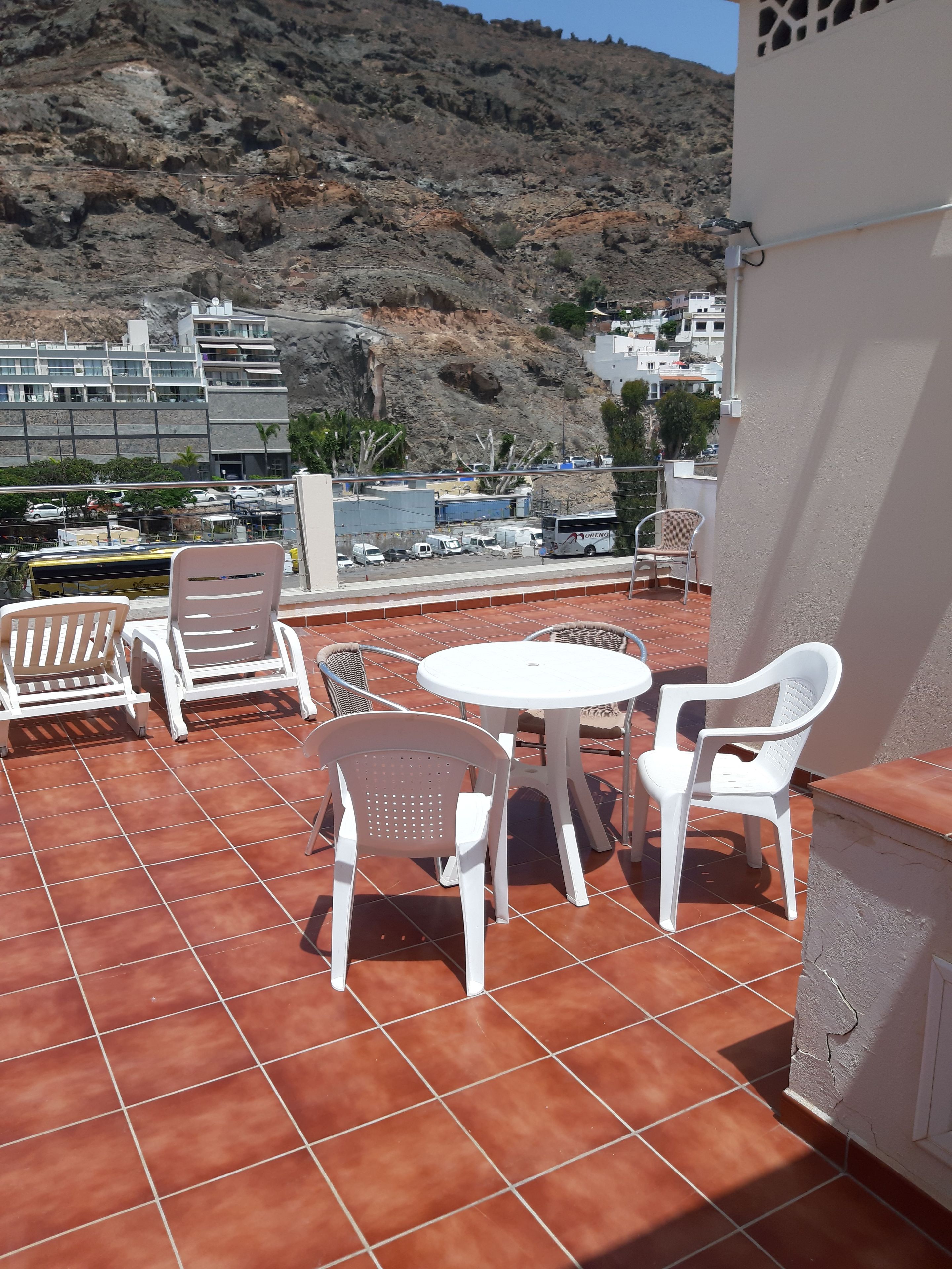 A terrace with white plastic chairs and a table, overlooking a rocky hillside and buildings.