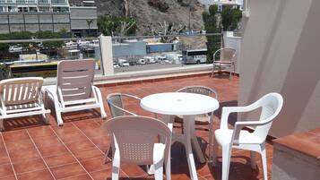 A terrace with white plastic chairs and a table, overlooking a rocky hillside and buildings.