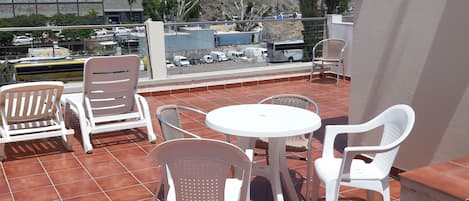 A terrace with white plastic chairs and a table, overlooking a rocky hillside and buildings.