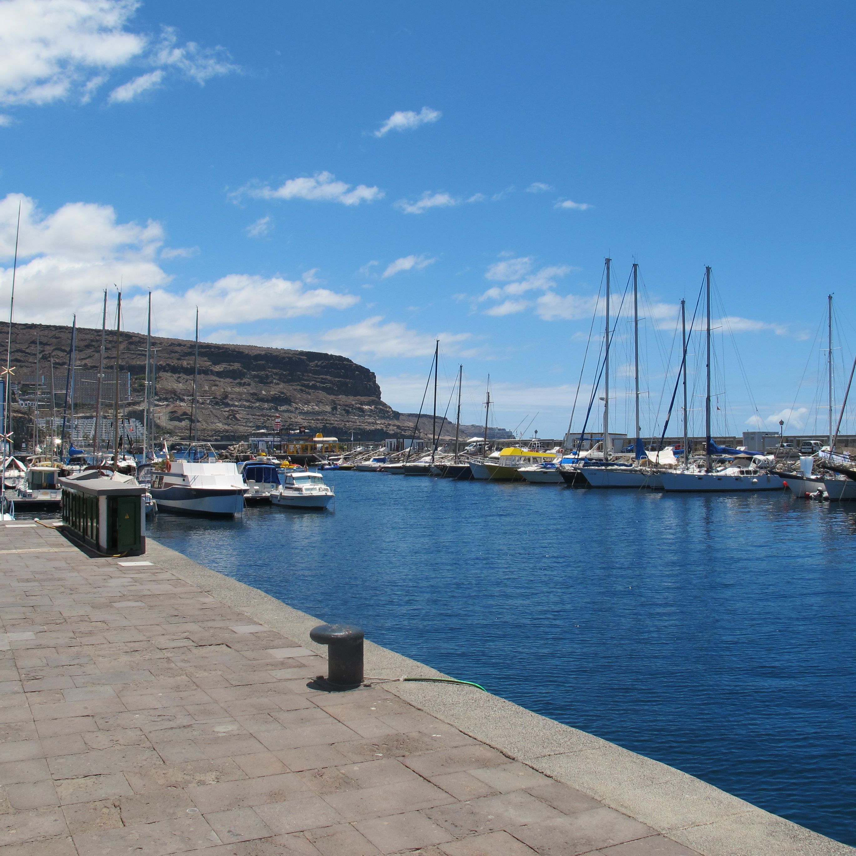 A marina with boats docked, a stone-paved walkway, and a rocky hillside in the background.