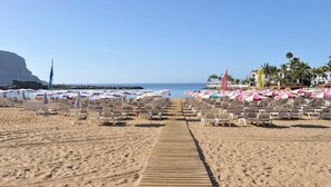 Una playa con filas de tumbonas y sombrillas, un paseo marítimo de madera y una vista despejada del mar y un acantilado al fondo.