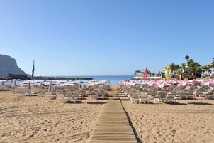 Una playa con filas de tumbonas y sombrillas, un paseo marítimo de madera y una vista despejada del mar y un acantilado al fondo.