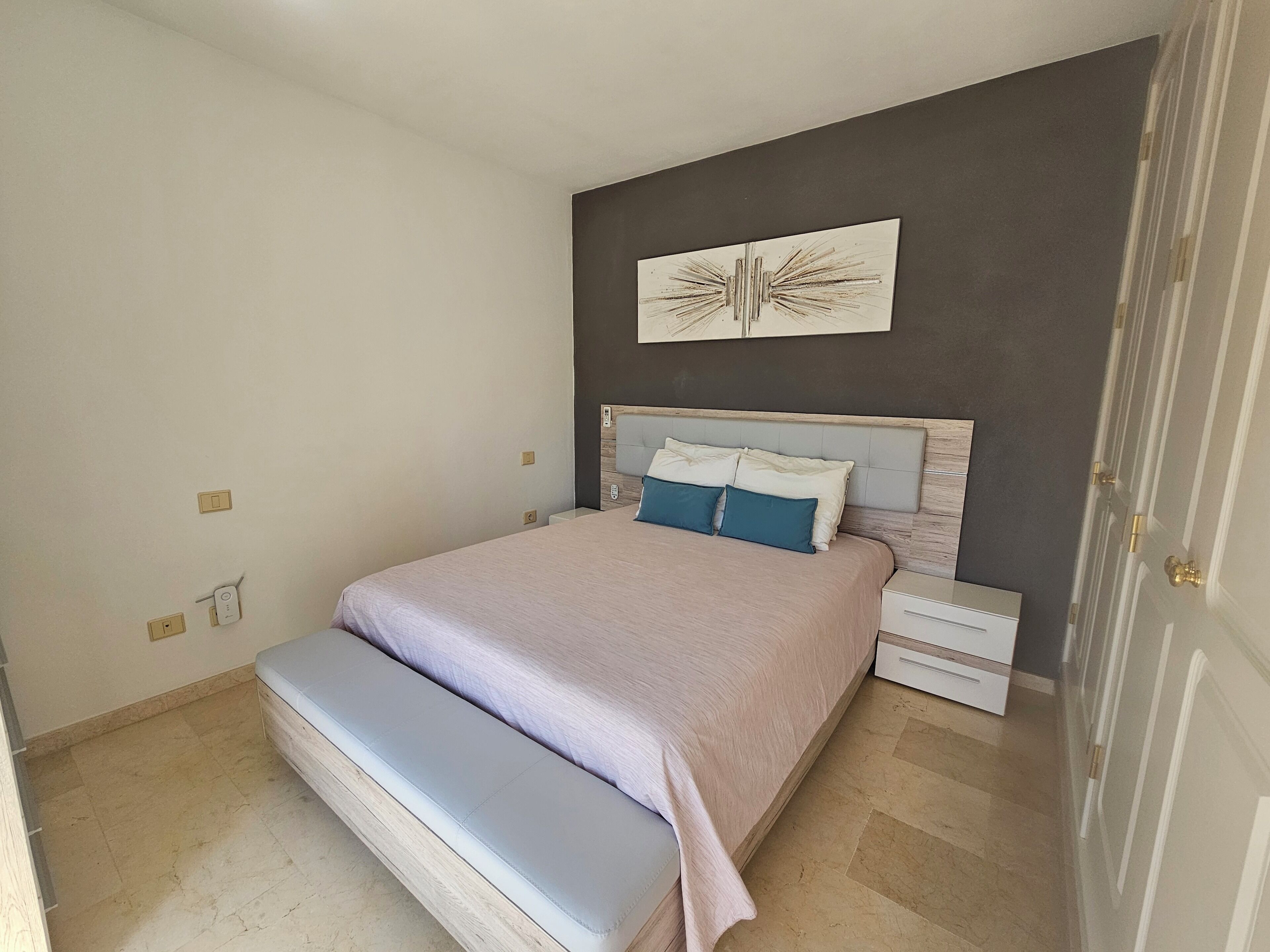 A bedroom with a bed, a bench, a nightstand, a wall-mounted artwork, and a door.