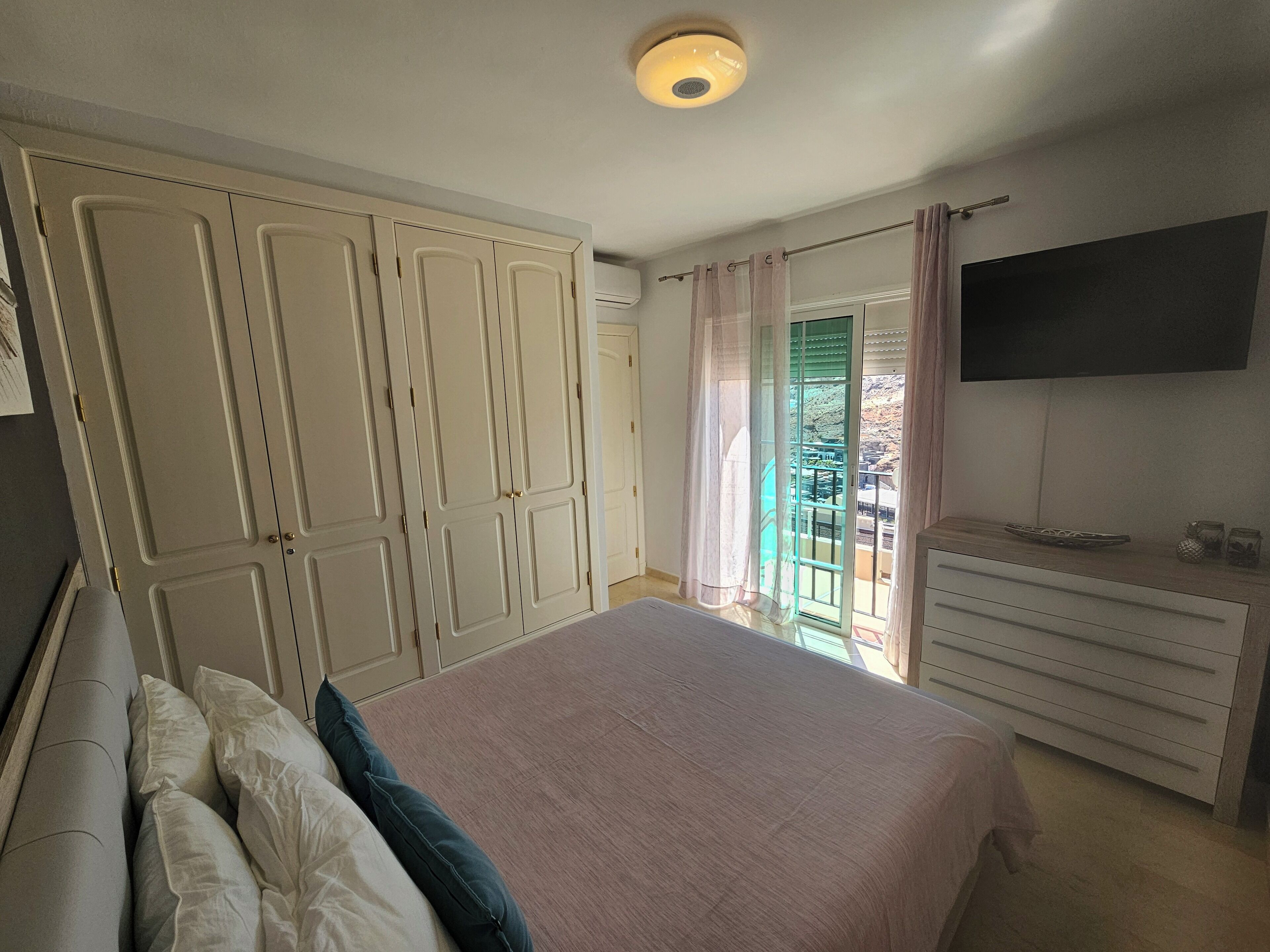 A bedroom with a bed, a dresser, a television, and a sliding glass door leading to a balcony.