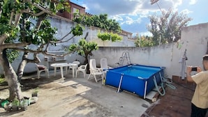 Un giardino con una piscina azzurra, sedie di plastica bianca, un piccolo tavolo e una scala che conduce alla piscina.