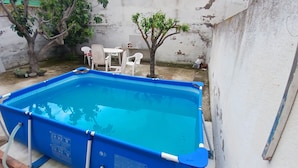 Una piscina rettangolare con interno blu, circondata da un muro bianco. Sullo sfondo, ci sono sedie in plastica bianca e un piccolo albero.