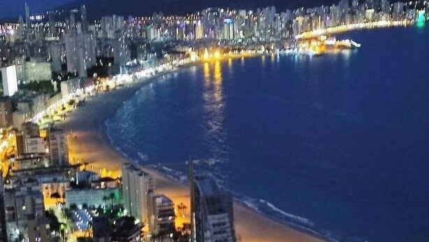 Eine beleuchtete Küstenstadt bei Nacht mit Gebäuden, einem Strand und einem Jachthafen.