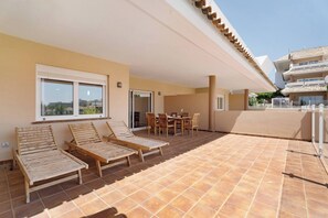 A sunlit outdoor area with two wooden lounge chairs and a table, a tiled floor, and a building with a balcony in the background. - Apto Royal Marina Golf V (San Miguel de Abona)