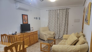 Een woonkamer met een houten eettafel, een beige bank, een houten salontafel, een televisie op een houten kast, en een groot raam met bloemen gordijnen.