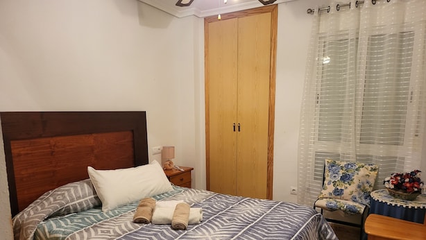 Een net opgemaakt bed met beddengoed met dessin, een houten garderobe, een stoel met een bloemenkussen en een klein tafeltje met een mand vol bloemen.