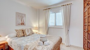 Een opgemaakt bed met een bedsprei met een patroon, een nachtkastje met een lamp, en een raam met gordijnen.