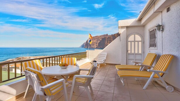 Een balkon met geel gestreepte stoelen en een tafel, met uitzicht op de kust, een rotsachtige klif en een heldere lucht.