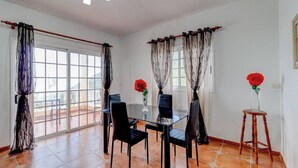A dining area with a glass table, black chairs, and a wooden stool. There are two windows with curtains, and a red rose in a vase on the windowsill. - Beautiful Arianny House With Pool (Arona)