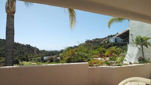 Ein Balkon mit einem beigen Sofa, einem kleinen Tisch und Blick auf einen angelegten Garten mit Palmen und einer Hügelkulisse im Hintergrund.