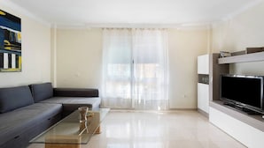 A modern living room with a sofa, a glass coffee table, a flat-screen TV, and a large window with sheer curtains. - Las Brisas Playa San Juan (Guia de Isora)