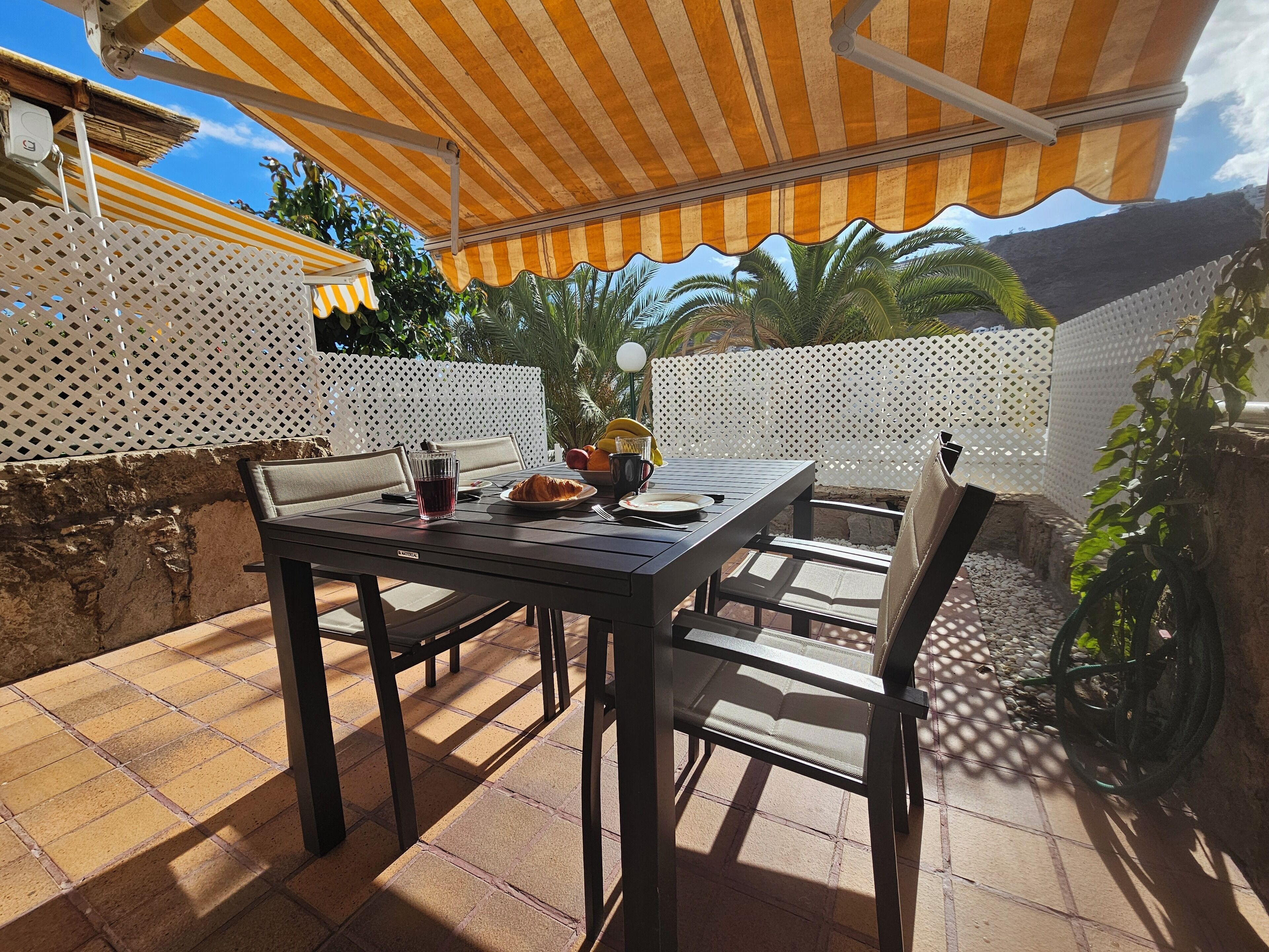 Un patio al aire libre con una mesa dispuesta para dos, un toldo a rayas arriba y una vista de palmeras y una montaña al fondo.