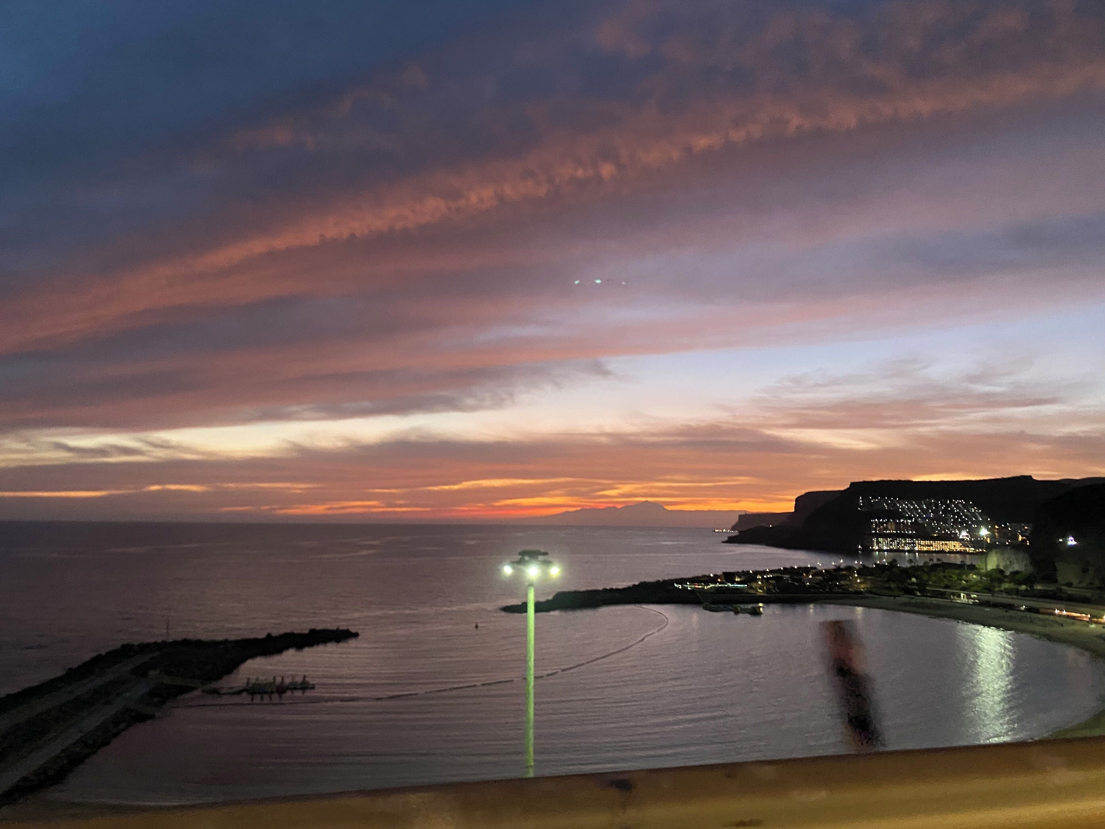 Vista de un atardecer costero con un mar tranquilo, un muelle y un pueblo lejano iluminado.