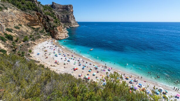 Una playa con aguas turquesas cristalinas, acantilados rocosos y numerosas sombrillas.