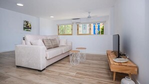 Una sala moderna con un sofá color crema, dos mesitas redondas, una mesita de noche de madera y un televisor.