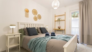 Une chambre à coucher comprenant un lit, des tables de chevet, un luminaire suspendu, une fenêtre avec des rideaux et une petite étagère sur laquelle est posée une plante.