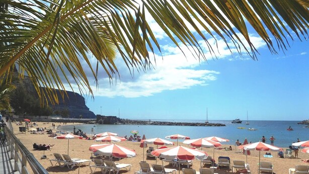 A beach with rows of sun loungers and umbrellas, a rocky coastline, and boats in the water. - Playa De Mogan Mirador 5 by El Sirocco (Mogan)