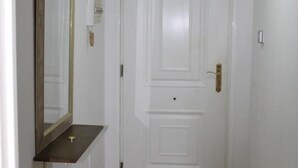 Um corredor com uma porta branca, um espelho à esquerda e um aparelho de iluminação no teto.