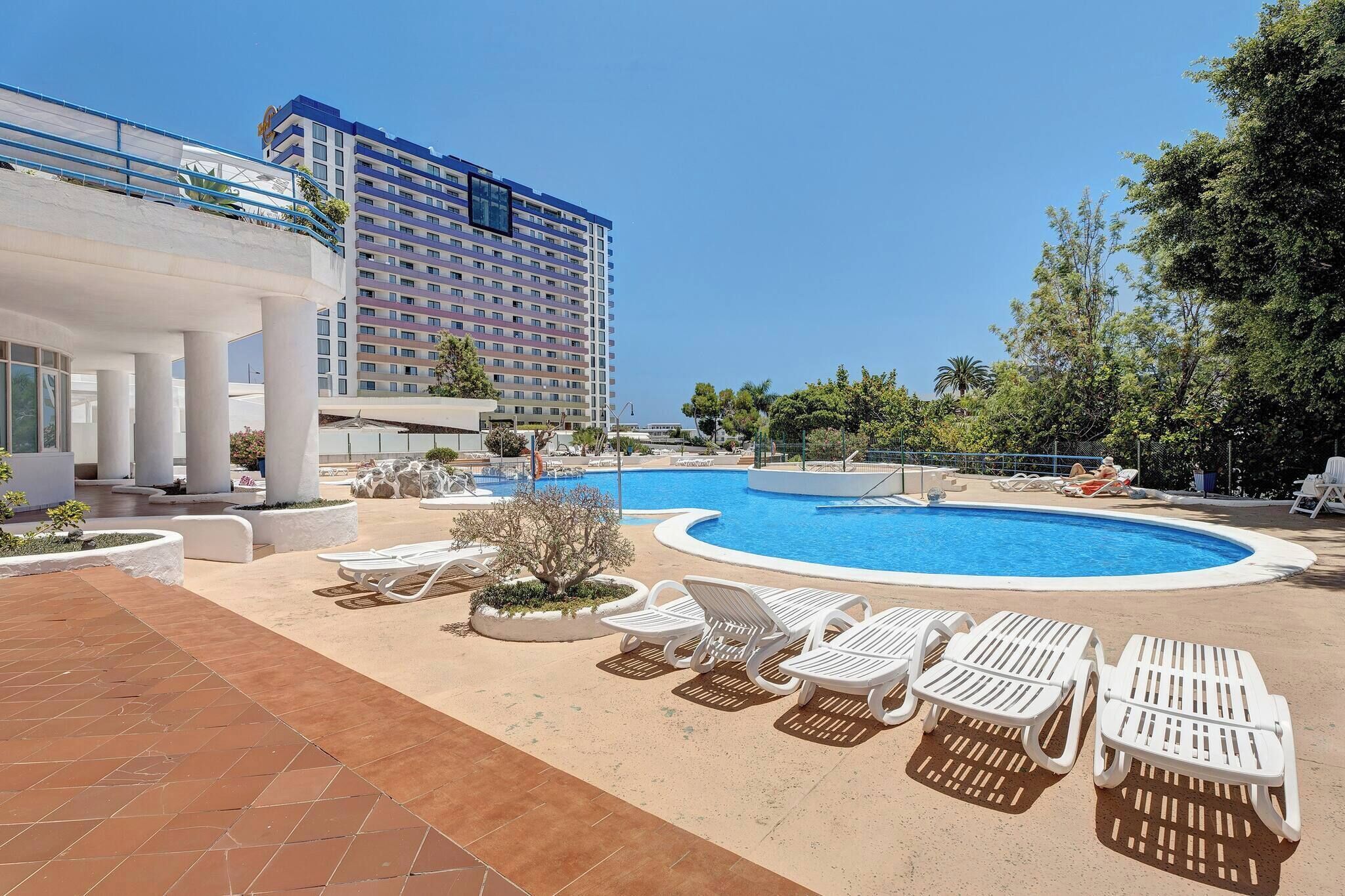 A hotel pool area with sun loungers, a large swimming pool, and a modern building in the background.