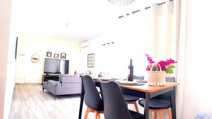 A modern living room with a grey sofa, a dining table with black chairs, a flat-screen TV, and a potted plant with pink flowers. - Villa Descansa With Private Pool, Air-conditioning, Sea View, Smart-tv & Wifi (Yaiza)