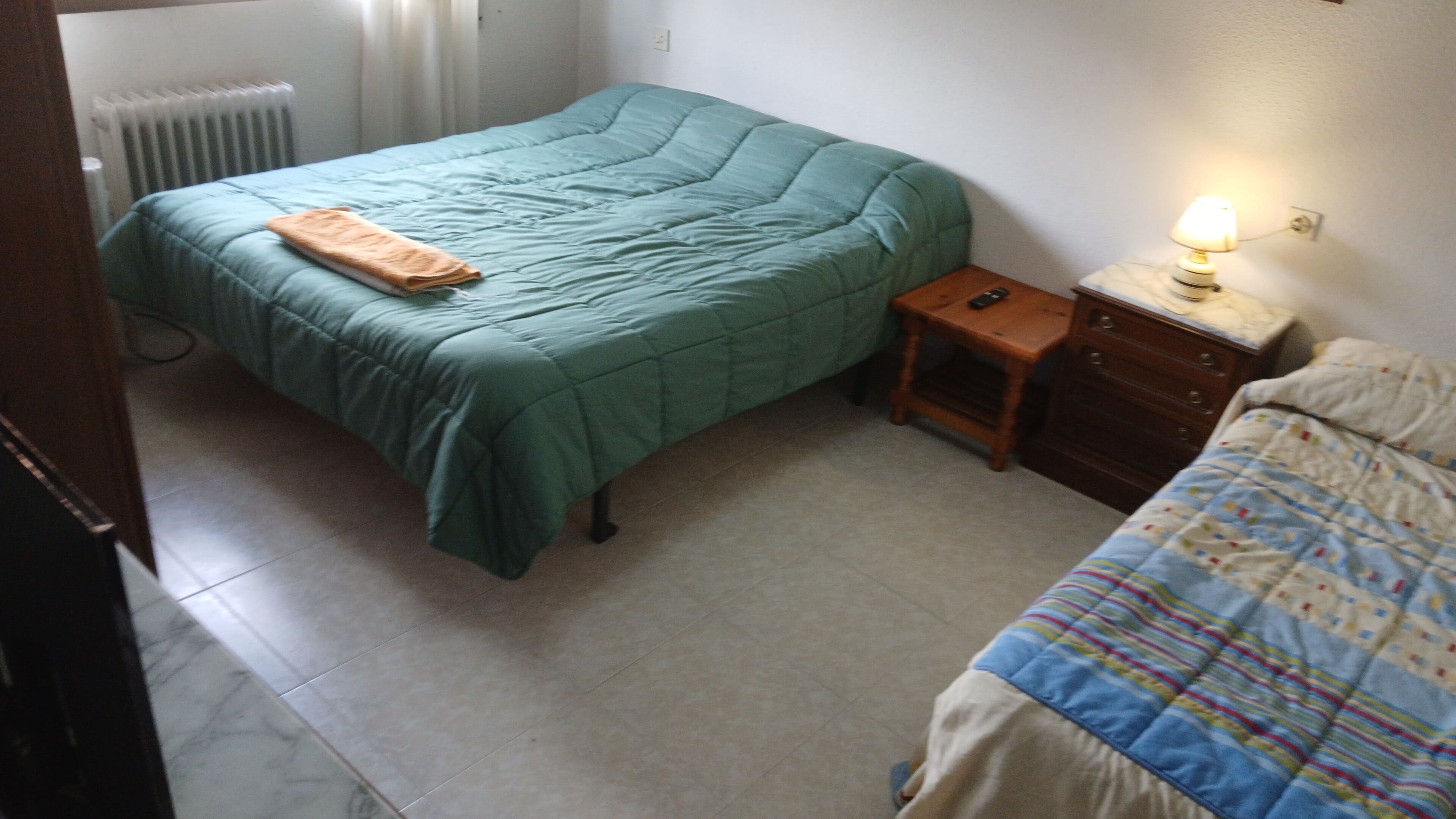 A bedroom with a green bed, a smaller bed with a patterned bedspread, a nightstand with a lamp, and a radiator.