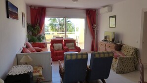 A living room with a sofa set, a coffee table, and a TV on a wooden cabinet. There is a window with red curtains and a view of palm trees outside. - Beautiful 2 Bedroom, 2 Bathroom Apartment (Roquetas de Mar)
