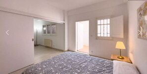 A bedroom with a bed, a nightstand with a lamp, a window with blinds, and a door leading to another room. - Beautiful Studio in Sant Gervasio (Barcelona)