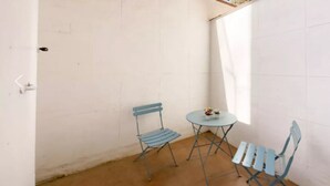 Een klein, afgesloten patio met een vloer en muren van witte tegels, met twee blauwe metalen stoelen en een kleine ronde tafel met een bloevas.