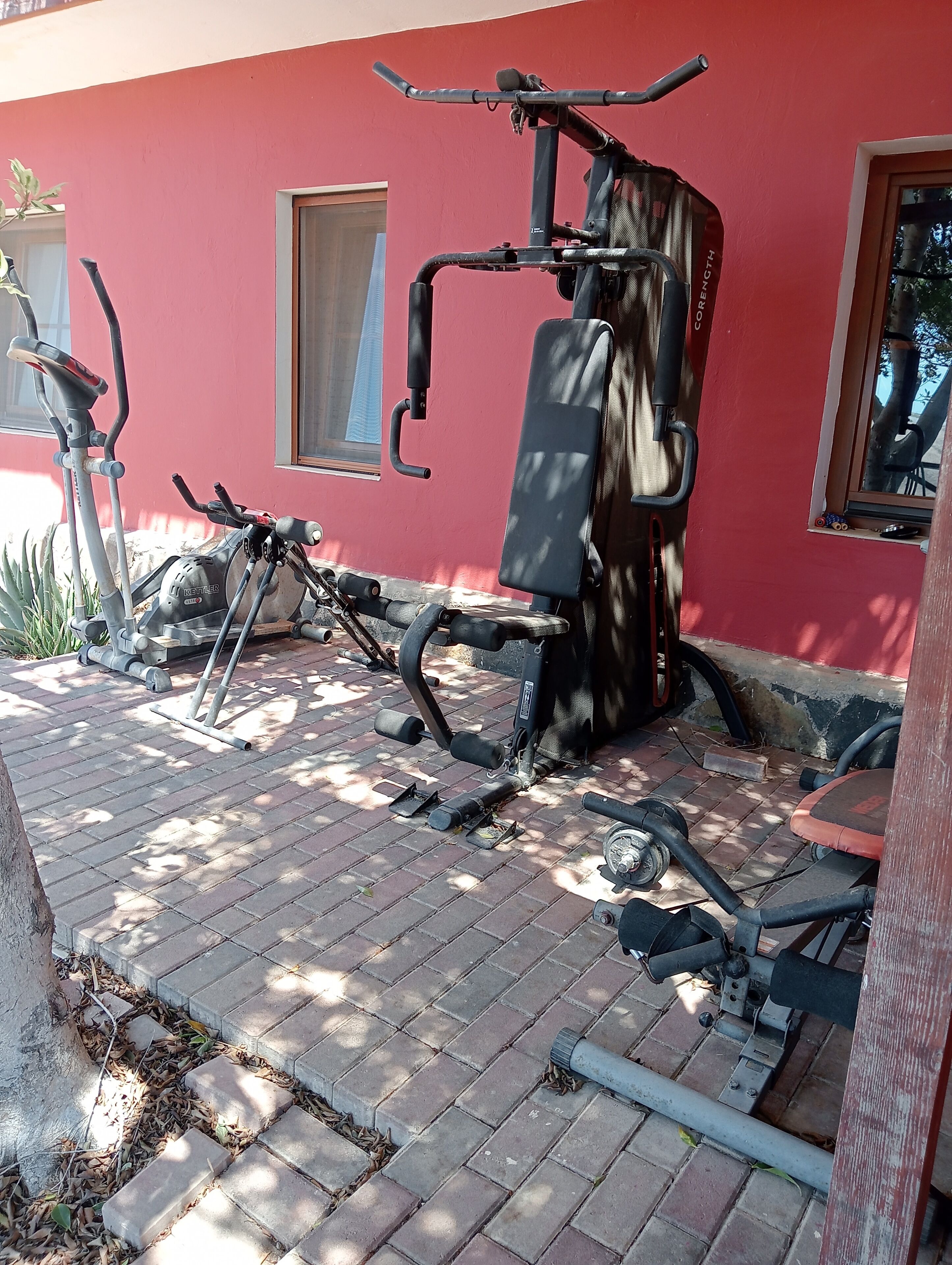 Un espace d’exercice en plein air comprenant un vélo d’appartement, un banc et un appareil de musculation, le tout adossé à un mur rouge.
