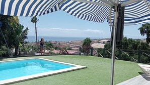 Area piscina con vista sull'oceano, ombrellone a righe e sedie a sdraio.