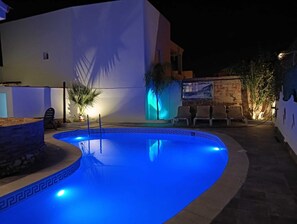 Une piscine dont le bord est illuminé de lumière bleue, entourée d’un bâtiment blanc avec un mur de pierre et des plantes en pot.