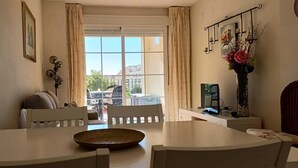 A dining area with a white table and chairs, a window with curtains, a view of buildings outside, and a vase with flowers on a side table. - Lovely 2 Bedroom, 2 Bathroom Apartment With Pool (Roquetas de Mar)