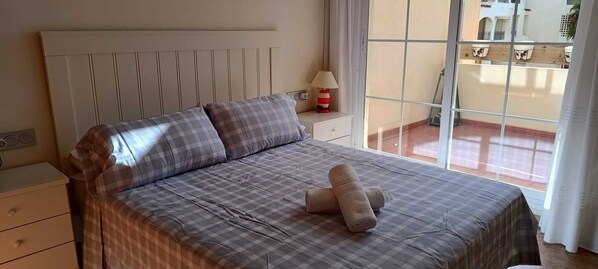 A neatly made bed with checkered bedding, a bedside lamp, and a view of the outdoor balcony through a window.