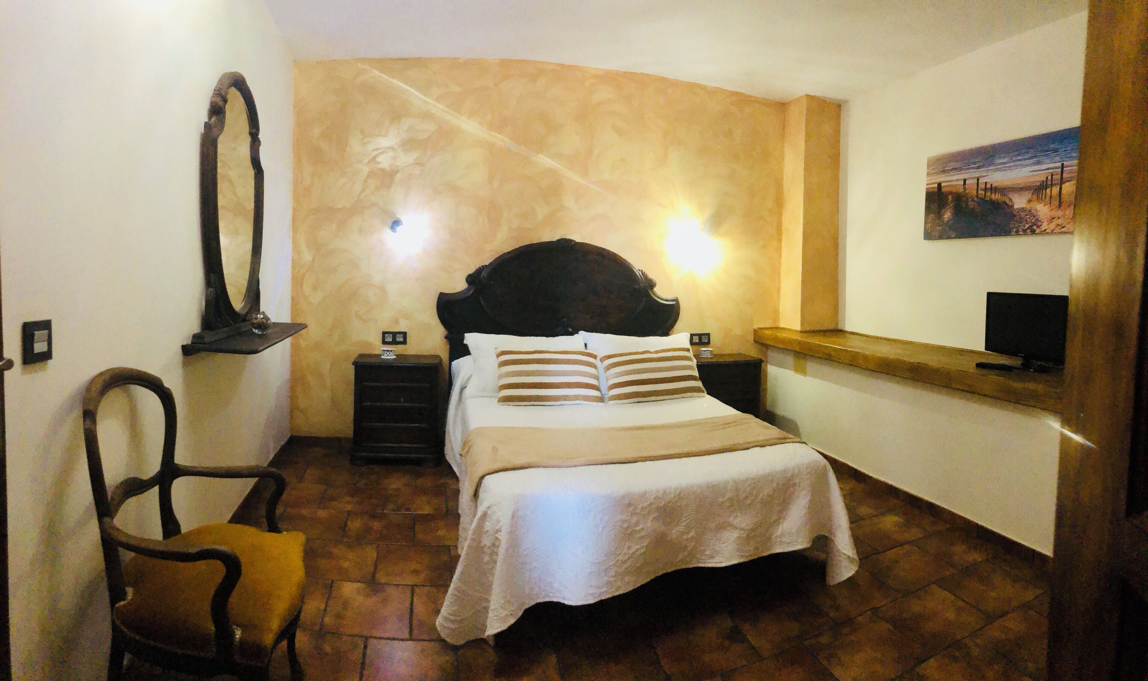 A bedroom with a curved wall, a bed with striped pillows, a wooden headboard, a nightstand, a mirror, a chair, and a painting on the wall.