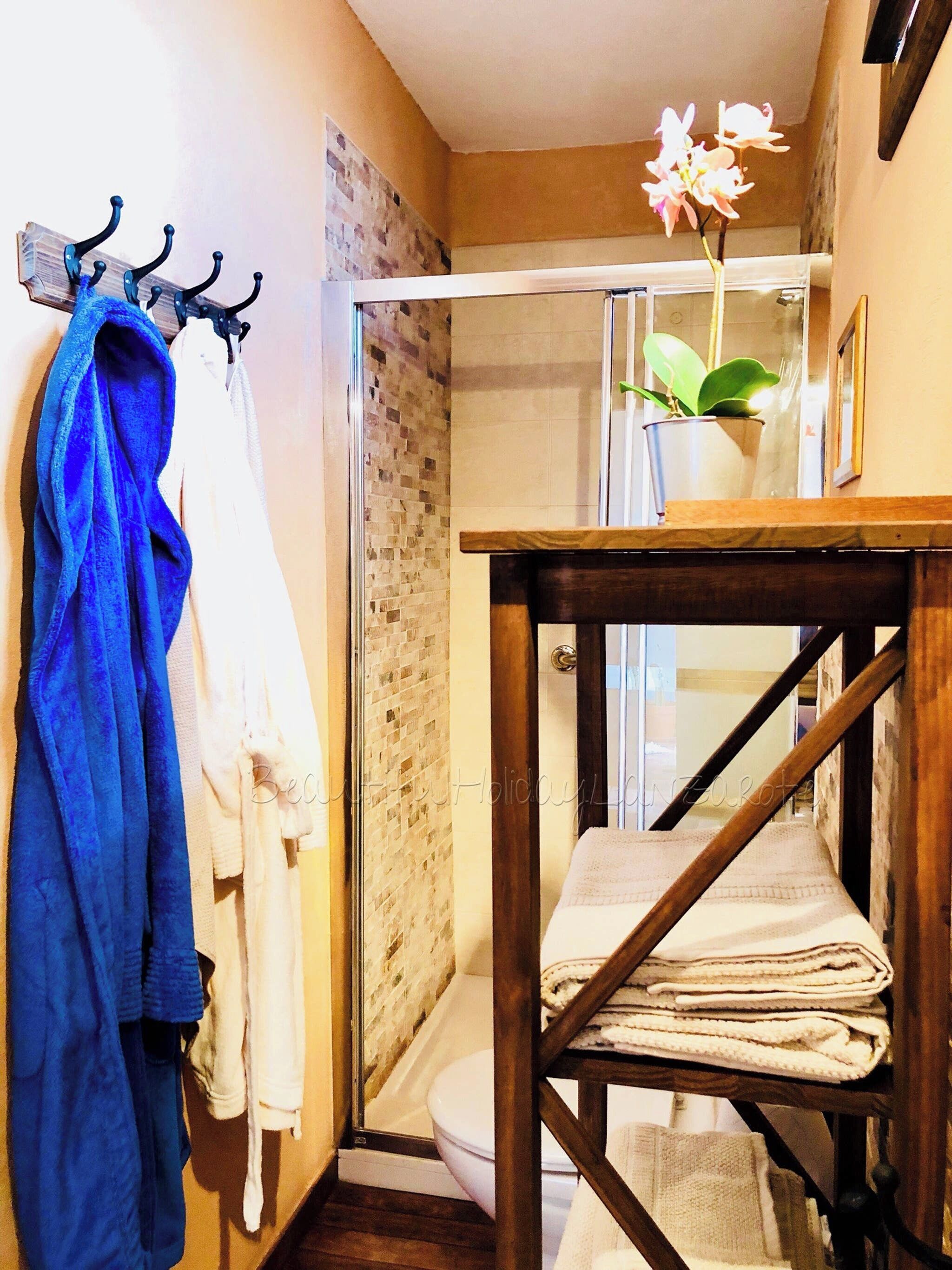 A bathroom with a glass shower, a wooden table with towels, and a blue robe hanging on a hook.