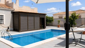 A swimming pool with a tiled deck, a covered area with a table and chairs, and a building with a closed shutter. - Townhouse With Shared Pool (Torrevieja)