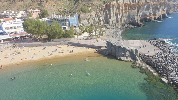 An aerial view of a coastal area with a sandy beach, rocky shoreline, and buildings in the background. - Mirador Studio Apartment (Mogan)
