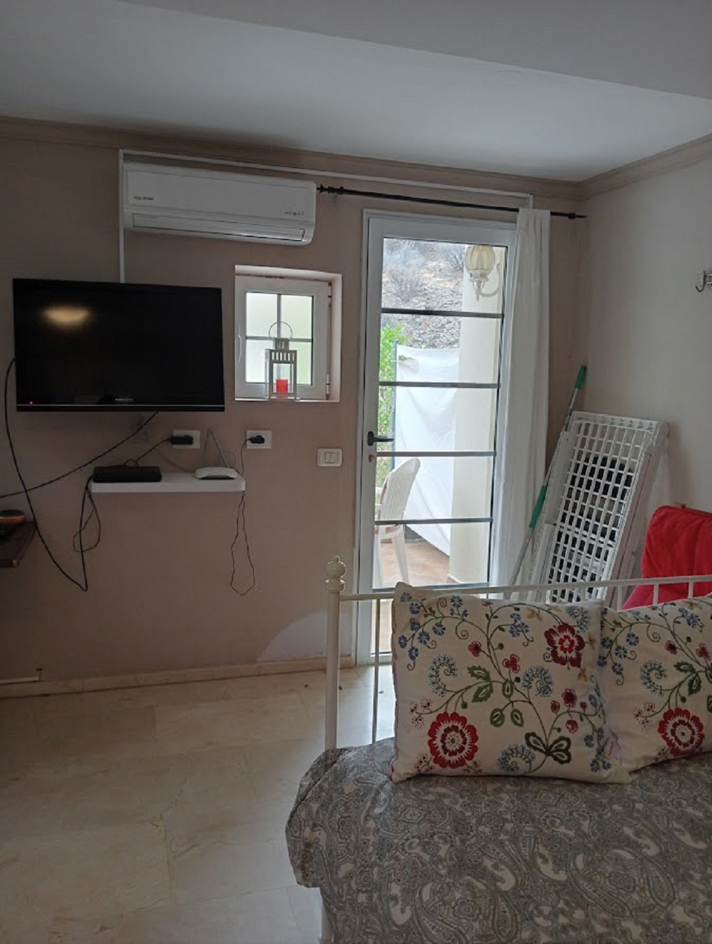 Una habitación con televisión, un ventanal pequeño, una puerta y una silla blanca con cojín estampado.
